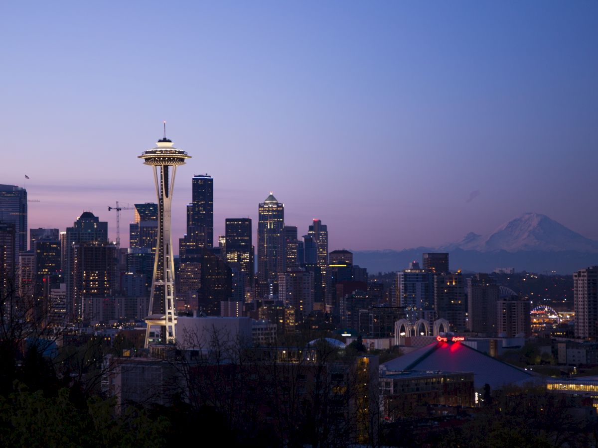 A twilight view of the Seattle skyline, featuring the Space Needle, with Mount Rainier visible in the background under a clear evening sky.