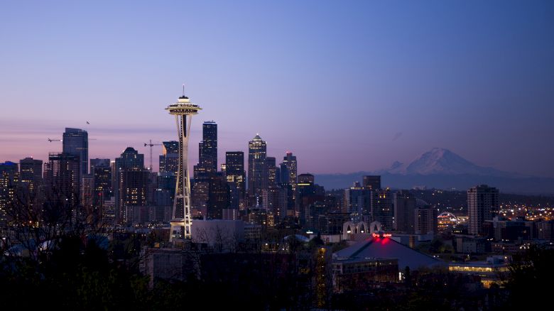 A twilight view of the Seattle skyline, featuring the Space Needle, with Mount Rainier visible in the background under a clear evening sky.