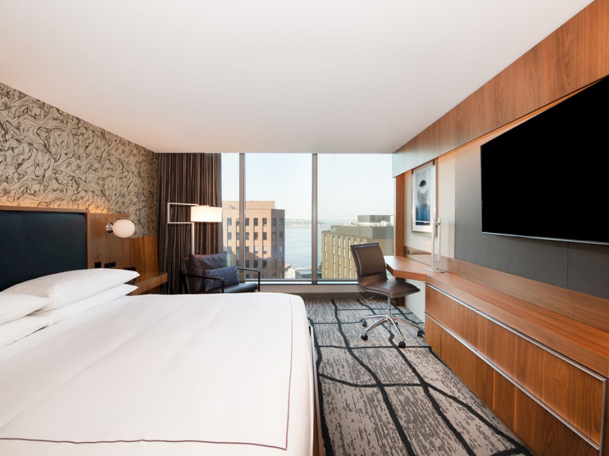 A modern hotel room with a king-size bed, large TV, desk, chair, and window with a cityscape view, featuring sleek decor and stylish furnishings.
