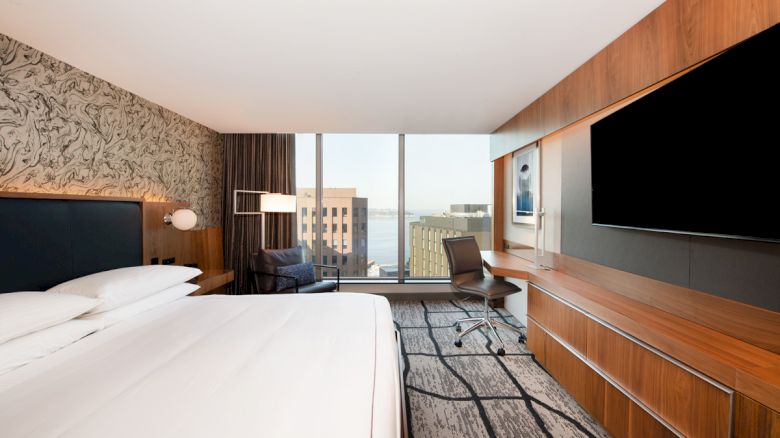 A modern hotel room with a king-size bed, large TV, desk, chair, and window with a cityscape view, featuring sleek decor and stylish furnishings.