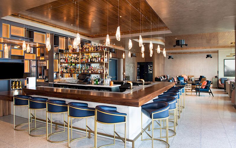 A modern, elegant bar with a large selection of alcohol, blue barstools, pendant lighting, and a lounge area in the background.
