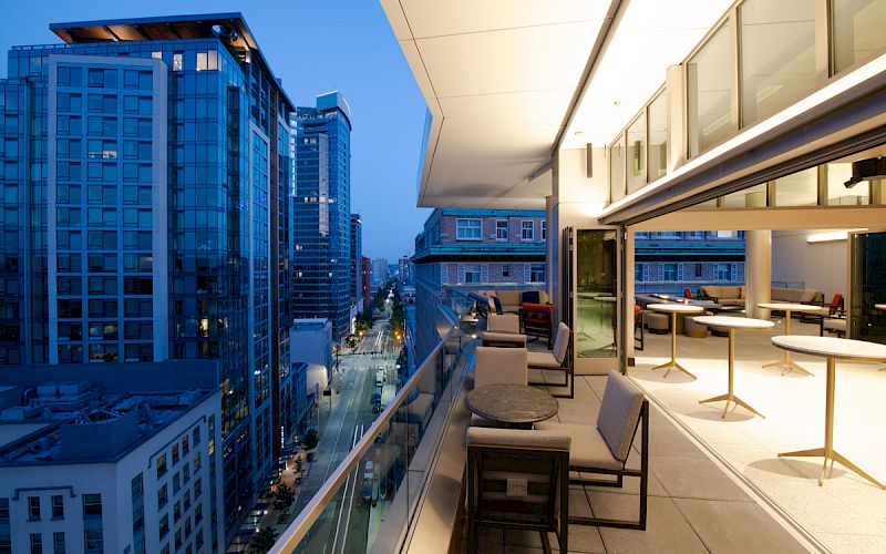 A modern outdoor patio with seating overlooks a city street lined with tall buildings at dusk. The space features tables, chairs, and minimal decor.