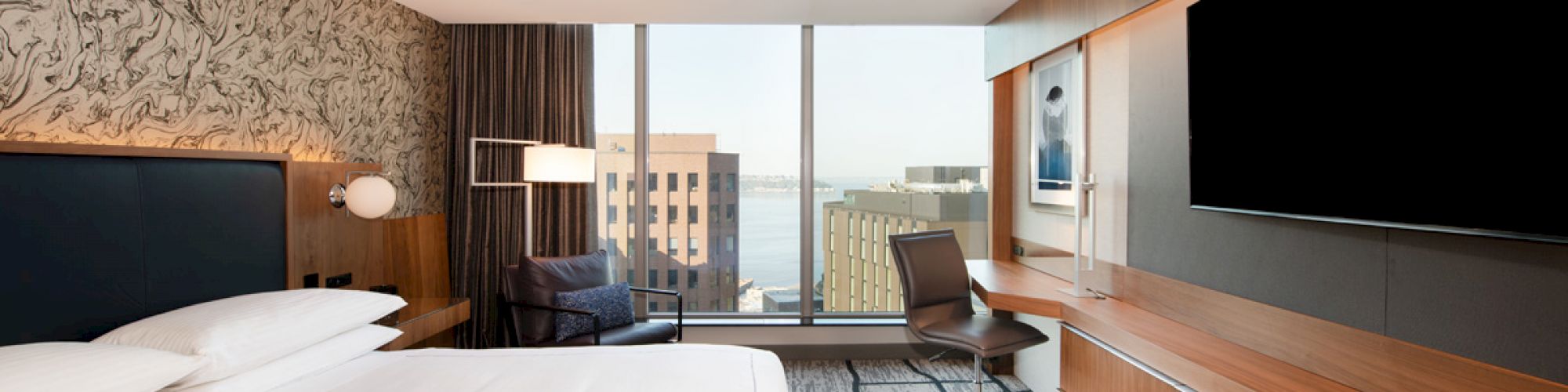 Modern hotel room with a large bed, flat-screen TV, desk, and a chair. Large window showcasing a city view with buildings and water.