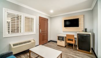 A small room with a table, chair, TV, mini-fridge, and microwave; it has a door, window with blinds, and an air conditioner below the window.