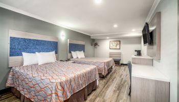 The image shows a modern hotel room with two double beds, a TV, a desk, and minimalistic decor. The room has wooden flooring and a neutral color palette.