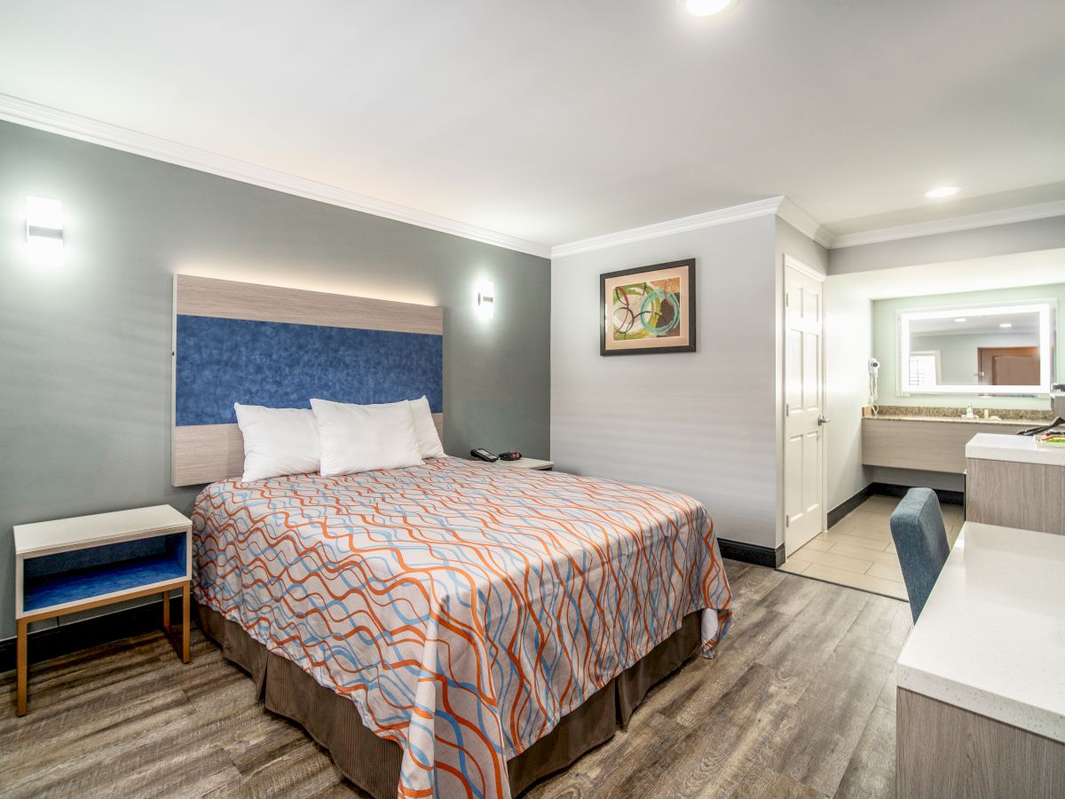 The image shows a modern hotel room with a bed, bedside table, wall art, desk, and a vanity area with a sink and mirror.