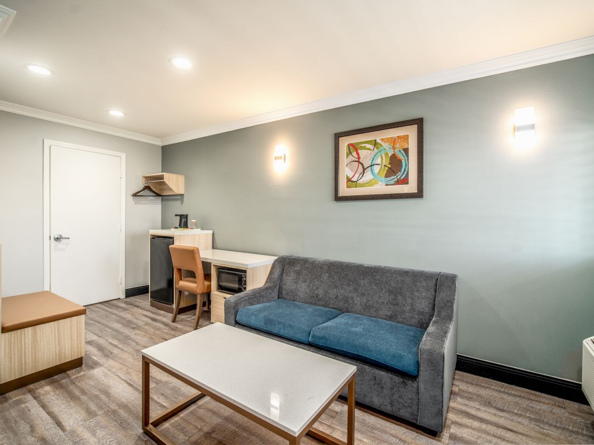 A modern, minimalist hotel room with a gray couch, a table, a desk and chair, coat rack, and a wall-mounted artwork, lit by soft lighting.