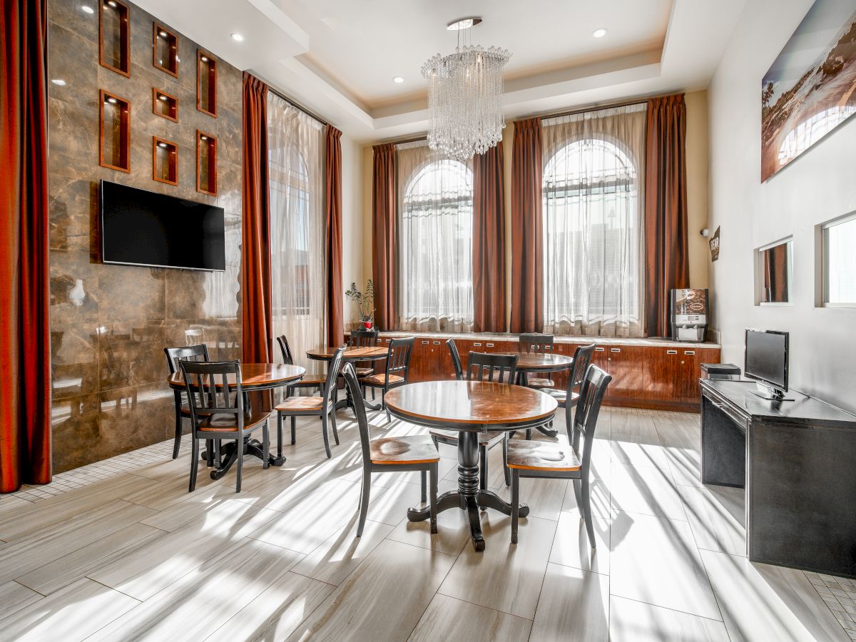 A modern, well-lit dining area with round tables, chairs, a large chandelier, wall-mounted TV, and tall windows with drapes.