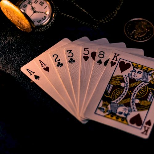 This image shows a hand of playing cards (two aces, 2, 3, 5, and a king), a pocket watch, and a coin, all on a dark surface.