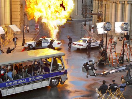 A film set at Universal Studios shows an explosion scene being recorded, with actors, a film crew, and an audience watching from a tour tram.