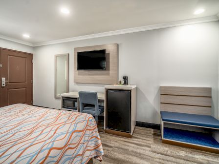 A hotel room featuring a bed, a wall-mounted TV, a desk with a chair, a small fridge, a coffee maker, a bench, and a door.