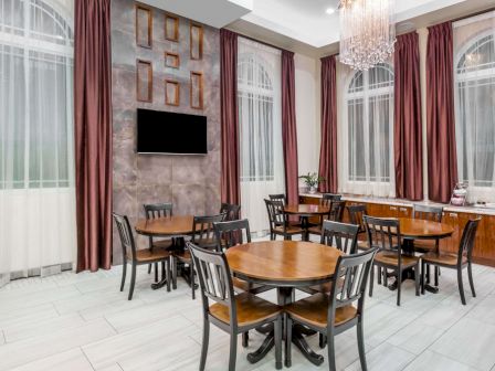 The image shows a dining area with several round tables and chairs, large windows with red curtains, a television, and a chandelier.