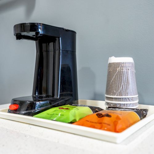 A coffee maker is placed on a white tray alongside stacked disposable cups and packets of coffee on a counter against a gray background.