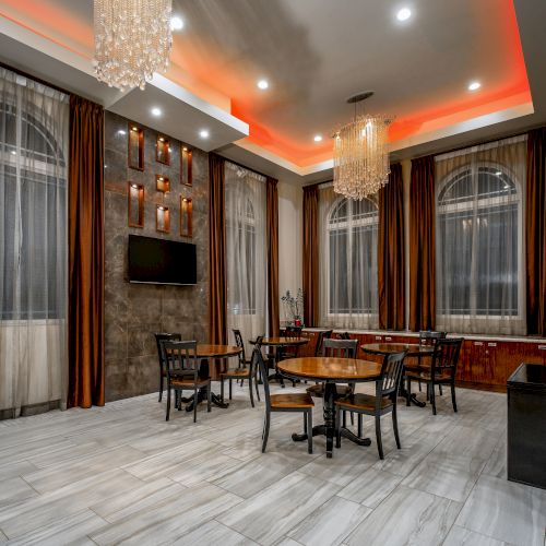 A modern room with round tables, chairs, chandeliers, tall windows with curtains, a TV, and magazine racks on a glossy tiled floor.